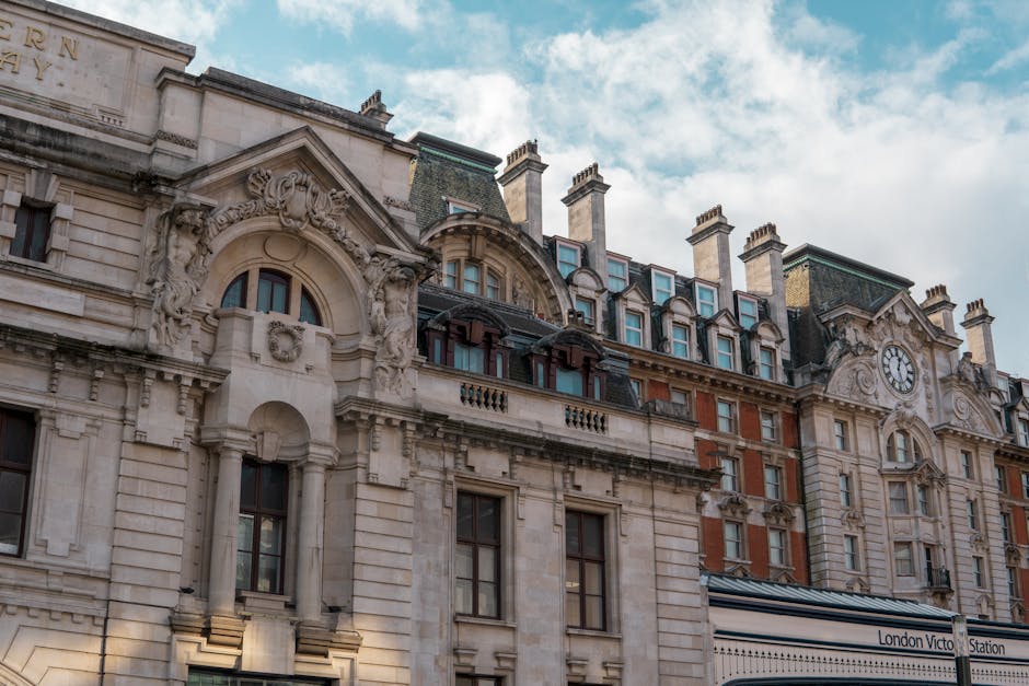 A view of Westminster Station in London with a large underground sign suspended from an ornate metal frame against a partly cloudy sky. Below, the entrance to the underground subway features a gated s